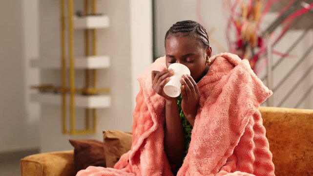 African woman wrapped in blanket drinking hot tea feeling sick with flu symptoms
