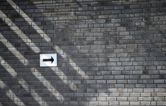 A dark grey brick wall featuring a white rectangular sign with a black arrow pointing to the right. Strong diagonal shadows cast across the left side of the wall.
