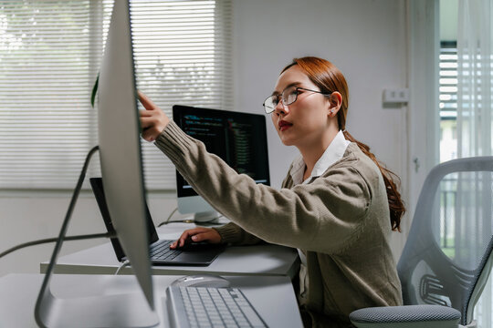 Young Asian woman programmer coding software, developing website on multiple screens from a home office workspace