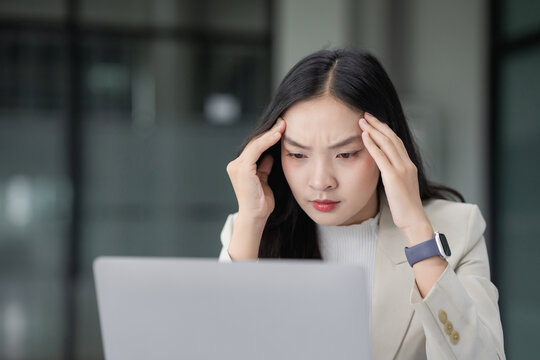 Young Asian woman feeling headache and stress while working on laptop in modern office, depicting burnout and fatigue