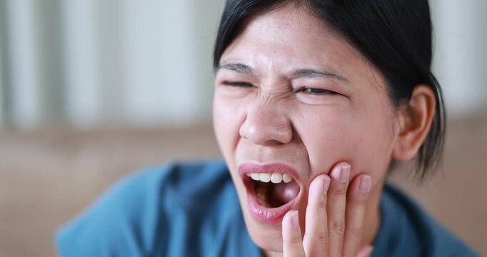 Woman suffering from toothache, holding her cheek in pain, tooth pain, dental problems, oral health, tooth sensitivity, cavity, dental treatment, and healthcare, close-up, Asian Thai people