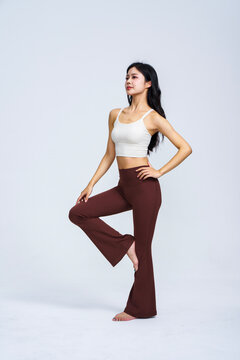 Young East Asian woman in white sports top and brown high-waisted yoga flare pants posing in plain light grey studio 