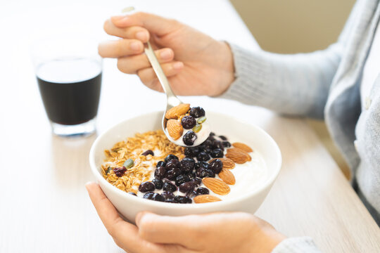 Close up of woman, girl  holding a bowl of healthy Greek yogurt topped with crunchy granola, fresh blueberries, and almonds, using a spoon to eat her breakfast with black coffee.