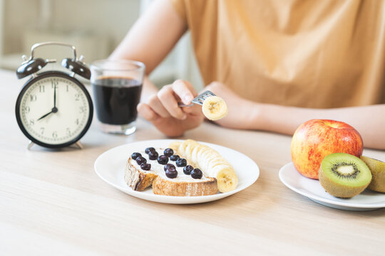 Young woman having a healthy breakfast with fruit toast and black coffee near an alarm clock showing eight AM, concept of morning routine and intermittent fasting diet lifestyle.