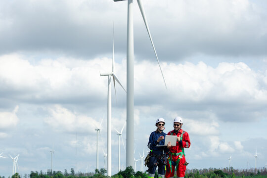 Two wind farm technicians use a laptop beneath large turbines while wearing safety gear, representing teamwork, renewable energy inspection, engineering, maintenance and clean electricity.