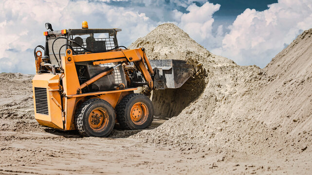 A construction machine is seen moving dirt and shaping a mound on a work site. A small compact loader moves sand for construction work