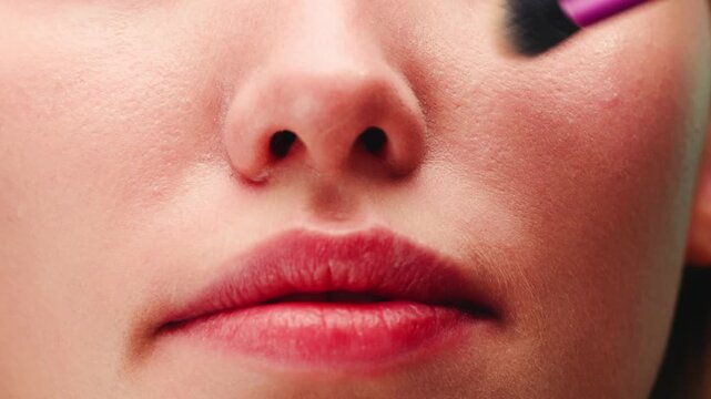Makeup artist applying highlighter with brush to young teenager student face, beauty close up on pink background, cosmetics advertising for youth