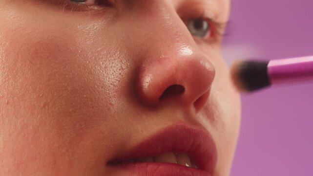 Makeup artist applying highlighter with brush to young woman face on pink background. Beauty close up for cosmetics, blush, powder and makeup advertising for youth.