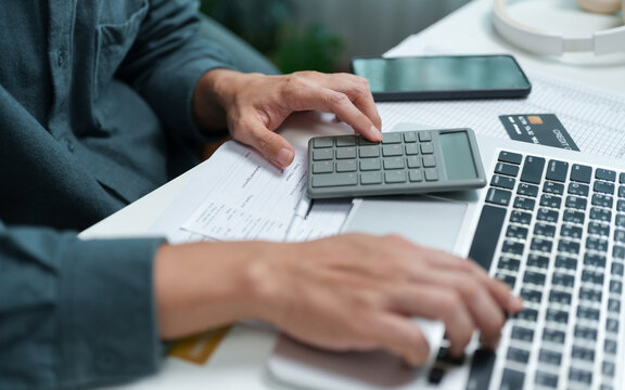 Man using calculator and laptop while reviewing bills and financial documents at home, representing debt management, credit payments, budgeting, expenses, loans and personal finance planning.