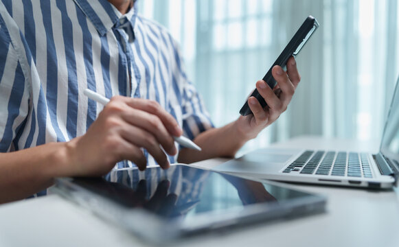 Male Freelance designer multitasking with smartphone, tablet, stylus and laptop at a desk, representing creative work, digital technology, productivity, mobility and modern workflow.