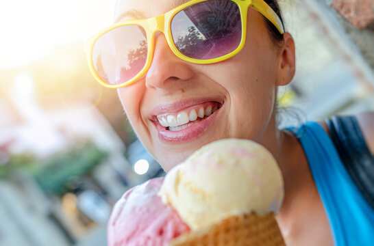 A cheerful woman wearing yellow sunglasses smiles while holding a waffle cone with two scoops of ice cream outdoors on a sunny day.