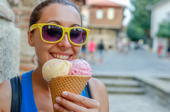 A cheerful young woman in yellow sunglasses enjoys a waffle cone with scoops of ice cream outdoors on a sunny summer day.