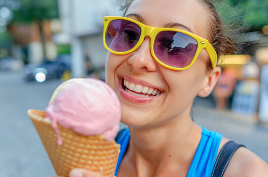 A cheerful young woman in yellow sunglasses smiles while holding a delicious pink ice cream cone outdoors on a sunny summer day.