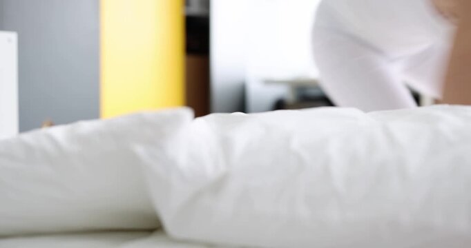 In a bright bedroom, a woman wakes in a rush, startled by an alarm. She fights against sleep, grappling with her pillows, and finally checks the time anxiously, desperate to start her day.