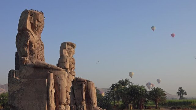 View of the Colossi of Memnon with hot air balloons floating in the sky above palm trees and desert landscape in Luxor Governorate, Egypt.