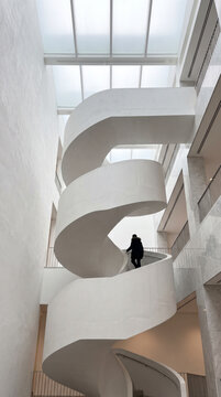 Solitary figure descends a sweeping white spiral staircase in misty atrium light