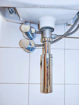 Chrome siphon and supply pipes under white ceramic washbasin. Under view of sink plumbing. Bathroom installation and plumbing maintenance concept. Close-up