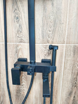 Black shower mixer faucet with control valves and handheld spray on wood-look tiled wall. Modern black bathroom fixtures. Shower system close-up. Bathroom renovation and plumbing concept