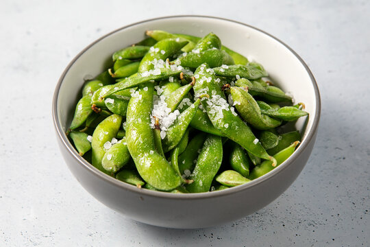 Fresh steamed edamame soybeans served in a bowl with coarse sea salt on a light textured background.
