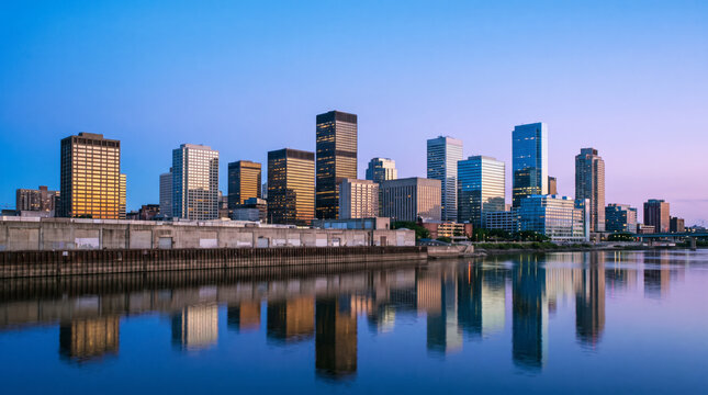 Riverfront financial district towers glow at dusk above a calm reflective waterway