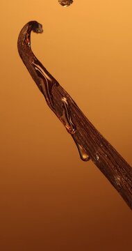 Macro vertical video of liquid oil flowing down a vanilla bean pod on amber background