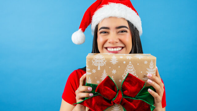 Smiling Latina woman wearing a santa hat and red shirt holding a wrapped gift box