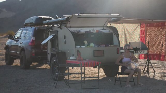 A mature woman enjoys playing a doumbek drum while sitting next to her teardrop camper in the Mojave desert at sunset.
