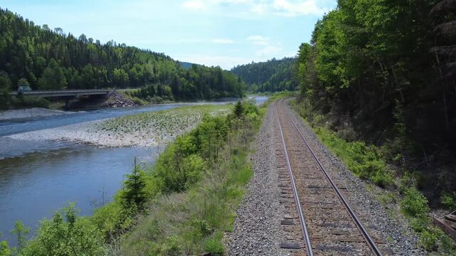 Drone footage documents rail transportation infrastructure running parallel to the Matapedia River in a scenic valley featuring a modern concrete roadway bridge.