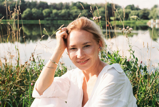 A woman smiles softly among tall grasses beside a still lake