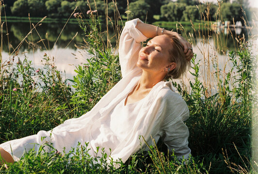 A woman rests in tall grass by a sunlit riverside