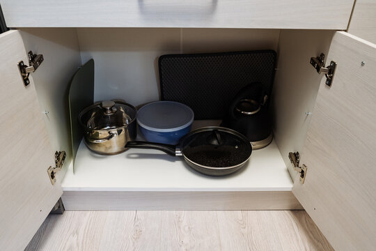 Open lower kitchen cabinet interior with two hinged doors and neatly arranged cookware on wooden shelf including frying pan pot kettle and storage container set