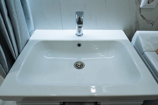 Modern bathroom sink basin with chrome faucet, round drain and marble tile backsplash in clean white countertop view, with folded shower curtain and toilet tank at edge