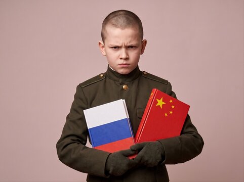 Sullen Slavic boy in cadet uniform holding Russian and Chinese flag books on red background. School education and language learning concept. Studio portrait for international education campaign