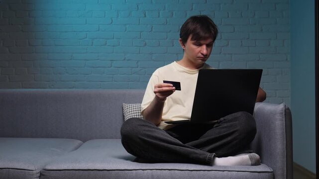 Young man on a sofa using a laptop and holding a credit card for online shopping in a modern living room with exposed brick, relaxed and secure payment