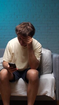 Vertical video young man sitting on a couch, holding his phone, feeling sad and depressed after reading an upsetting message or suffering from social media addiction and digital stress