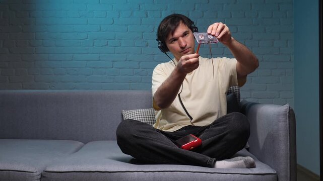 Young man in headphones sits on a sofa rewinding an audio cassette with a pencil, lost in nostalgia for retro analog music technology and memories of 80s90s listening