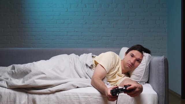 Man lying in bed under a blanket late at night, holding a game controller and staring at the screen, tired and bored from long gaming sessions and sleep deprivation