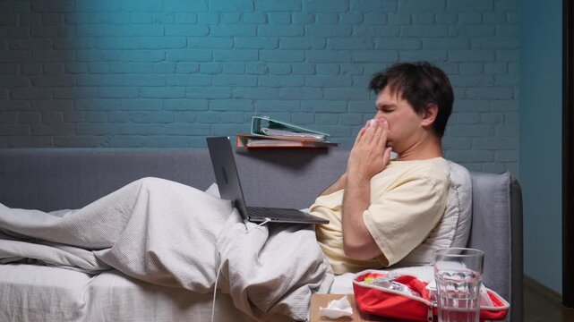 Young man lying in bed, feeling unwell and blowing his nose into a tissue, still trying to work on his laptop, experiencing illness and fatigue during remote work