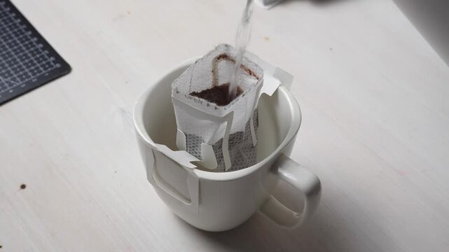 Close-up of pouring hot water into a drip coffee bag inside a white ceramic mug in 4K. Ground coffee brewing process with an alternative drip filter method on a light wooden table background