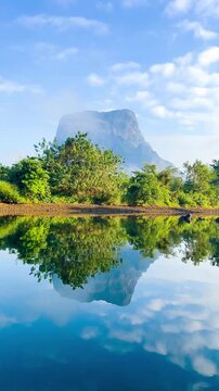 Landscape features the distinctive flat-topped mountains of Muang Fuang, Laos. Reflections of the scenery and a sky filled with fluffy clouds on the gently rippling water, set amidst lush greenery.