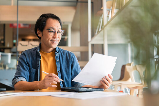 Focused Asian consultant reviewing business paperwork with content expression, analyzing financial report and strategic contract while planning project development in modern bright creative office