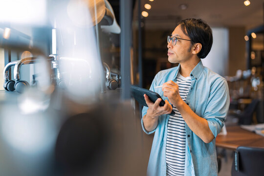 Focused Asian store supervisor using digital tablet to inspect showroom stock, auditing product quality and retail inventory management with professional diligence in modern furniture design store