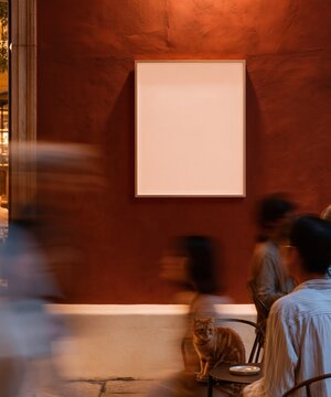 Tabby cat sitting on a chair by a blank poster mockup on a warm mocha brown stucco wall, featuring motion blur people for a cozy cafe atmosphere