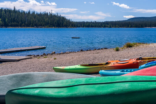 Kayaks and canoes rest on the pebbled shore of Elk Lake in Deschutes County, Oregon, with a dense pine forest in the background.