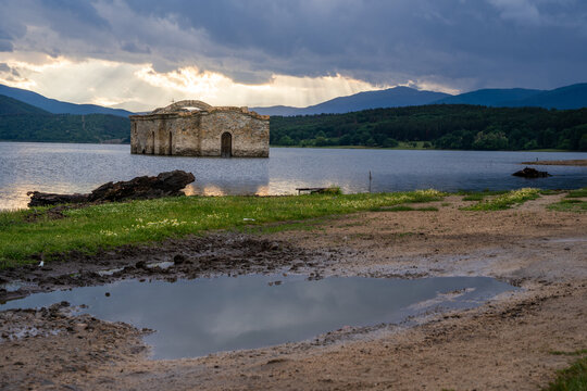 View of the submerged St. John of Rila church in a lake, with dramatic sunbeams breaking through storm clouds over distant mountains and a muddy shoreline in Sliven, Bulgaria.