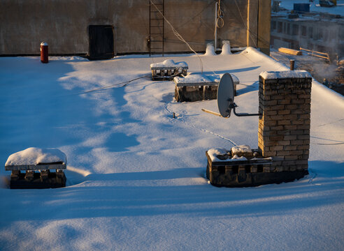 View of a snow-covered rooftop featuring a brick chimney, a satellite dish, and ventilation units under soft winter sunlight in Vilnius, Vilnius County, Lithuania.
