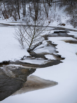 View of a partially frozen stream flowing through a snow-covered landscape with bare trees and ice formations in Vilnius, Vilnius County, Lithuania.