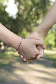 Vertical close-up of two people holding hands tightly outdoors against a soft blurred green park background. Interpersonal Support. Red String Protection