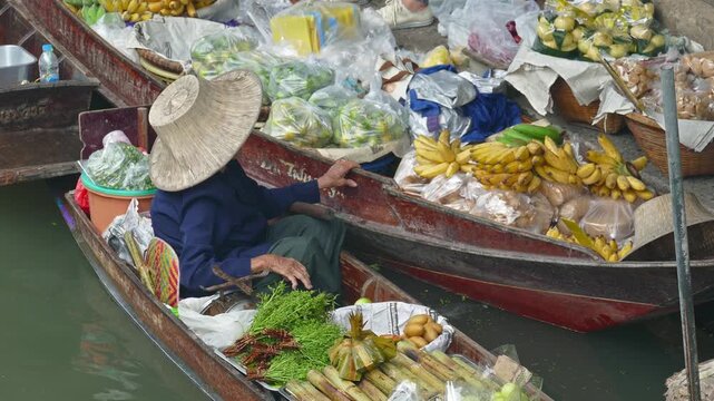Tambon Khunphithak, Chang Wat Ratchaburi, Thailand - 24 February 2017: View of a vendor in a traditional hat selling fresh produce from a wooden boat at the Floating Market.
