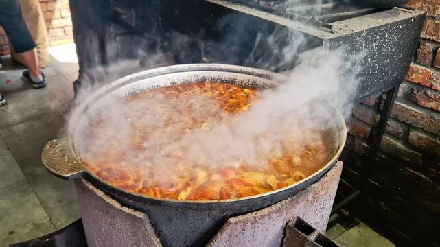 Hearty meat stew simmers vigorously in a cast iron cauldron with dense steam billowing outdoors. Authentic open fire cooking adds rich culinary texture to rustic food content.